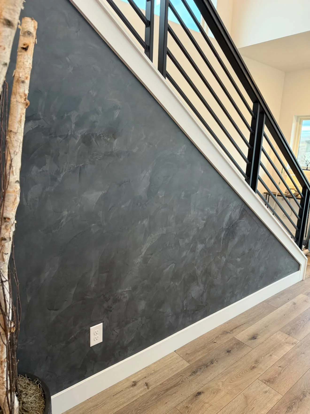 A near black Venetian plaster wall running up under a staircase, white oak floor, a black steel railing and birch branches